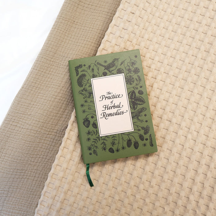 A green journal with hand-drawn illustrations of birds, flowers, and fruits in black, behind a white box of text that says The Practice of Herbal Remedies. A green ribbon sticks out of the bottom of the journal.