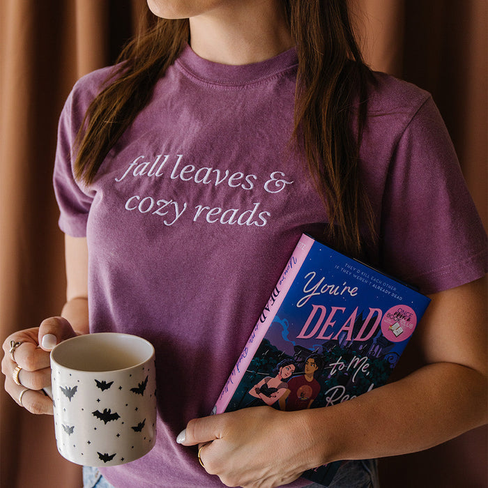 A woman wearing a purple graphic t-shirt with the text 'fall leaves & cozy reads' in white script. She is holding a white ceramic mug with black bat patterns and a book titled 'You're Dead to Me, Reed Walker'.