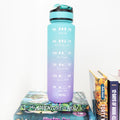A blue and purple ombre water bottle with goals to drink more water printed on the side sits atop a stack of books.