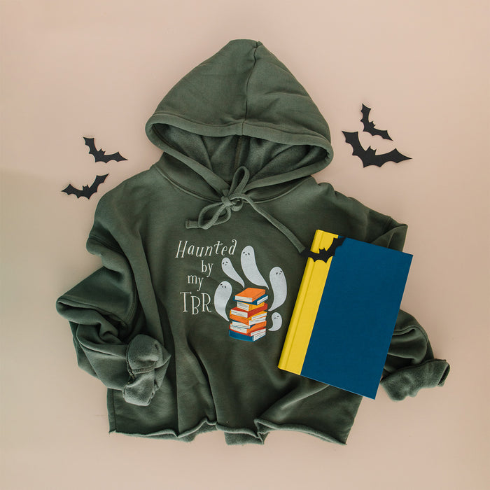 An olive-green hoodie with a 'Haunted by my TBR' graphic, styled in a flat lay with a blue and yellow notebook, black bat cutouts, and a beige background.