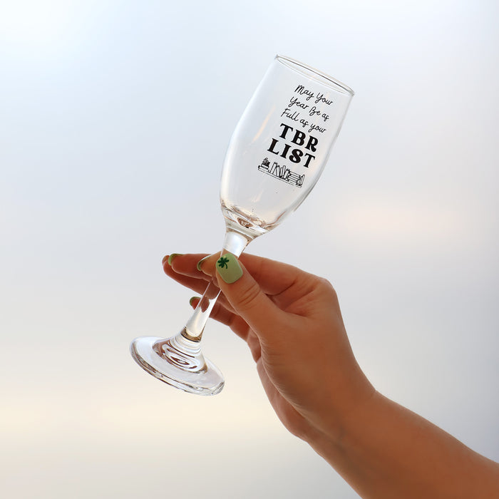 Hand holding a champagne flute that reads "May your year be as full as your TBR List" and features an etching of books along the bottom of the glass.