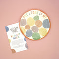 A multi-colored plate sits on a pink background. The plate says "Put a Fork in Me" along the top and has multiple colored dots on the plate labeled with numbers and symbols for various games. The instruction booklet sits next to the plate in the image.
