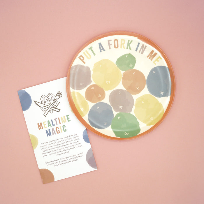 A multi-colored plate sits on a pink background. The plate says "Put a Fork in Me" along the top and has multiple colored dots on the plate labeled with numbers and symbols for various games. The instruction booklet sits next to the plate in the image.