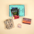A hardcover copy of Paradise Coast by Suzanne Young lays on a green OUABC box and is surrounded by three wrapped gifts labeled with page numbers.