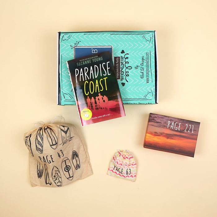 A hardcover copy of Paradise Coast by Suzanne Young lays on a green OUABC box and is surrounded by three wrapped gifts labeled with page numbers.
