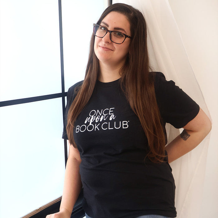 A woman with long brown hair and glasses wearing a black graphic t-shirt that reads 'Once Upon a Book Club' in white script, posing next to a window with white drapes.