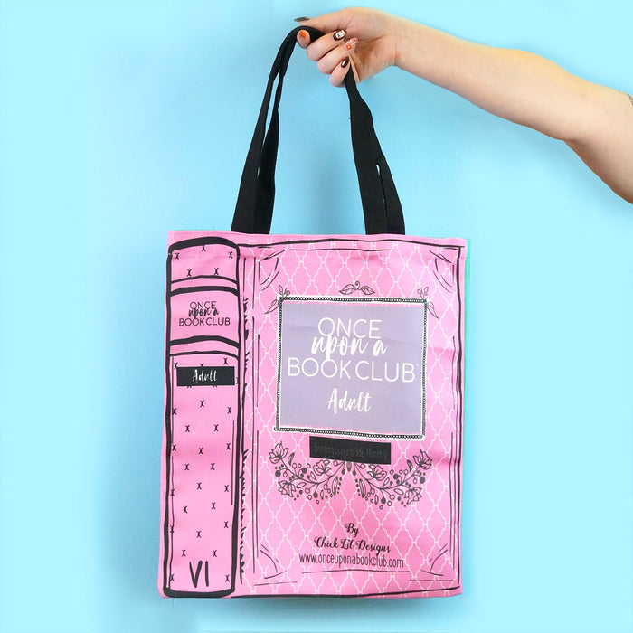 A hand holding a pink tote bag with a book-like design featuring the Once Upon a Book Club logo and the word "Adult" on the cover.