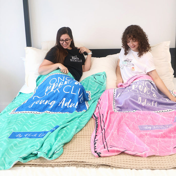 Two women sitting on a bed covered in colorful plush blankets. The blanket on the left is a turquoise and blue patterned throw blanket inspired by the Once Upon a Book Club Young Adult box and the blanket on the right is pink and purple, inspired by the Adult box.